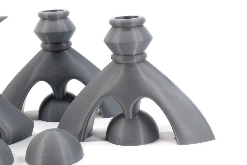 May include: Three gray, 3D printed candle holders with a unique, geometric design. Each holder has a round base and three curved arms that meet at the top to form a triangular opening. The holders are made of a smooth, matte material.