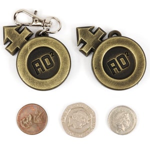 May include: Two gold-colored metal keychains with a black circle containing the text "AD^2" and a gold-colored arrow. Three coins are also shown in the image.
