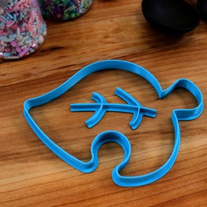 May include: A blue plastic cookie cutter shaped like a leaf with a smaller leaf design inside. The cutter is on a wooden surface.