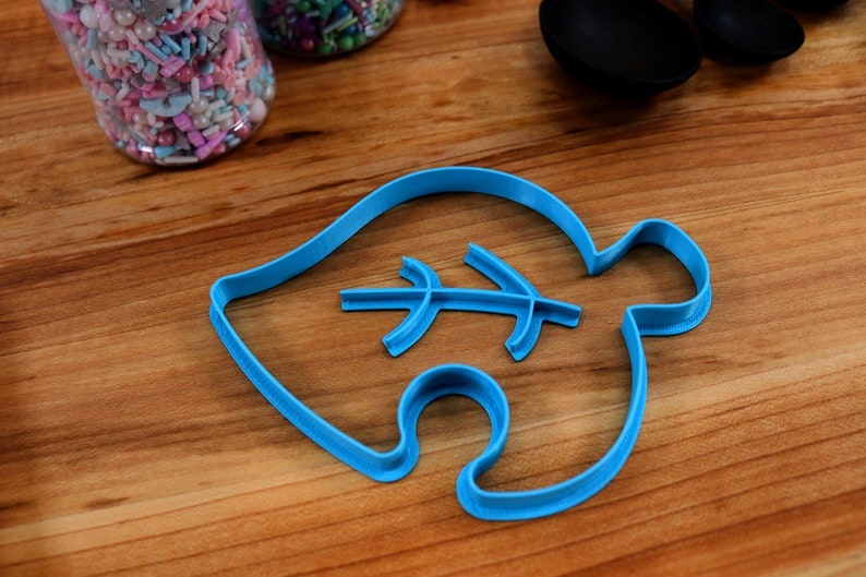 May include: A blue plastic cookie cutter shaped like a leaf with a smaller leaf design inside. The cutter is on a wooden surface.