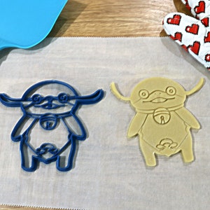 May include: Two cookie cutters in the shape of a cartoon character. One cutter is blue and the other is beige.