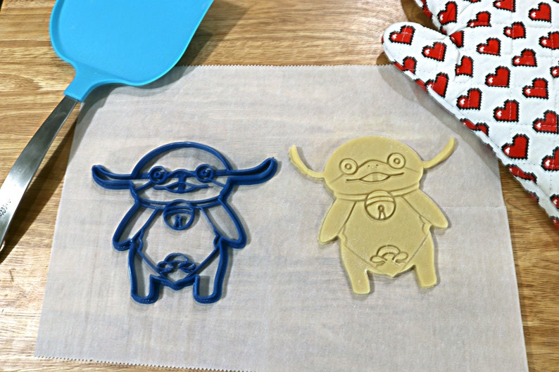 May include: Two cookie cutters in the shape of a cartoon character. One cutter is blue and the other is beige.