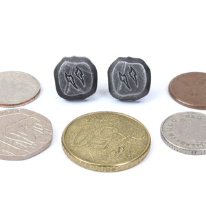 Puede incluir: Se muestran dos pendientes cuadrados de metal gris oscuro con un dise&ntilde;o estilizado. Varias monedas de diferentes tama&ntilde;os y colores rodean los pendientes, incluyendo una moneda de 25 centavos, una moneda de 50 peniques, una moneda de euro y un penique.