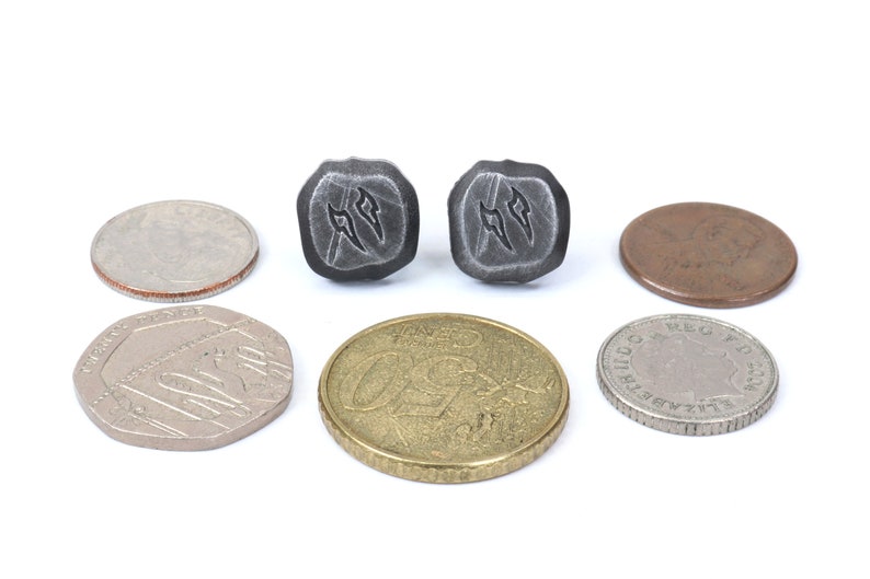 Puede incluir: Se muestran dos pendientes cuadrados de metal gris oscuro con un dise&ntilde;o estilizado. Varias monedas de diferentes tama&ntilde;os y colores rodean los pendientes, incluyendo una moneda de 25 centavos, una moneda de 50 peniques, una moneda de euro y un penique.