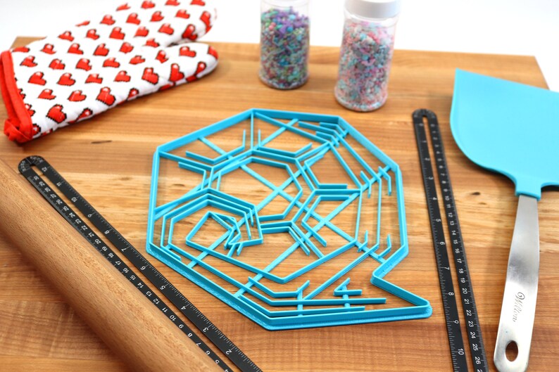 May include: A blue plastic snowflake cookie cutter on a wooden surface with a rolling pin, a ruler, a spatula, and two jars of sprinkles.