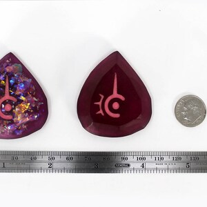 May include: Two teardrop-shaped pendants with a symbol, one with iridescent glitter and the other solid. A dime and a ruler are next to the pendants. The ruler measures up to 6 inches.