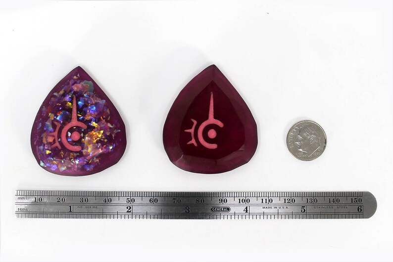 May include: Two teardrop-shaped pendants with a symbol, one with iridescent glitter and the other solid. A dime and a ruler are next to the pendants. The ruler measures up to 6 inches.