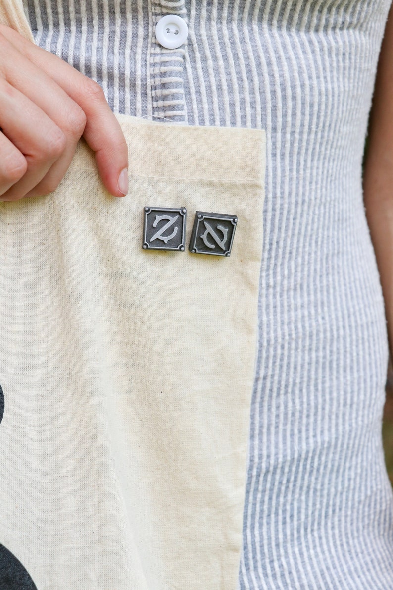 Op de afbeelding: Twee zilveren metalen pinnen met een gestileerd letter Z-ontwerp. De pinnen zijn bevestigd aan een witte tote bag.