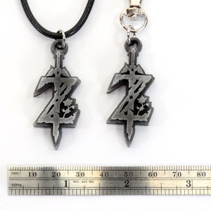May include: Two silver pendants featuring a stylized sword and a floral design. The pendant on the left is attached to a black cord necklace, while the pendant on the right is attached to a silver key chain. A ruler is visible in the background.