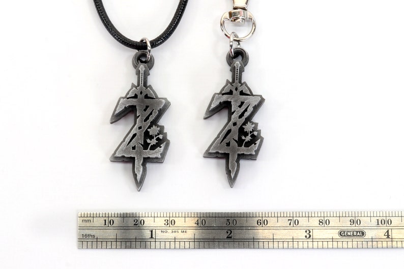 May include: Two silver pendants featuring a stylized sword and a floral design. The pendant on the left is attached to a black cord necklace, while the pendant on the right is attached to a silver key chain. A ruler is visible in the background.