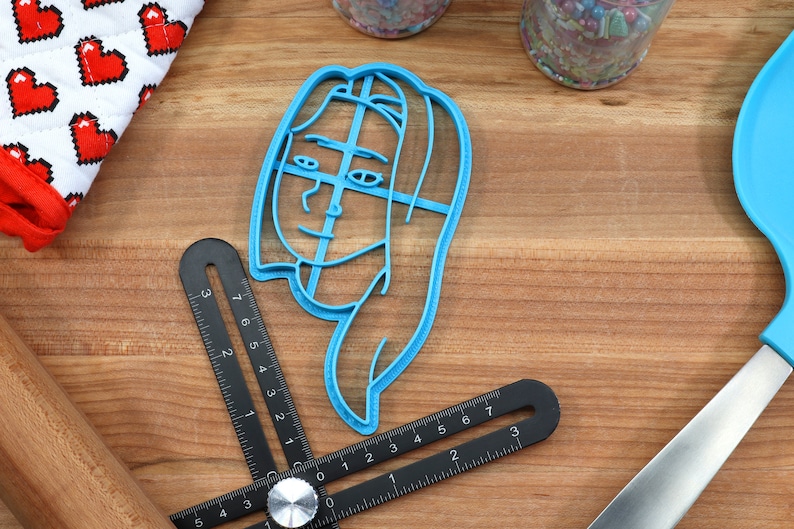 Op de afbeelding: Een blauwe plastic koekjesvormer in de vorm van een gezicht met een rasterpatroon op het gezicht.