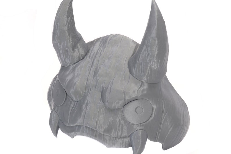 May include: A gray, textured, 3D printed mask with two horns and large, round eyes. The mask has a detailed, textured surface and appears to be made of a durable material.