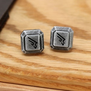 May include: Two silver cufflinks with a geometric design and a black symbol in the center. The cufflinks are on a wooden surface.