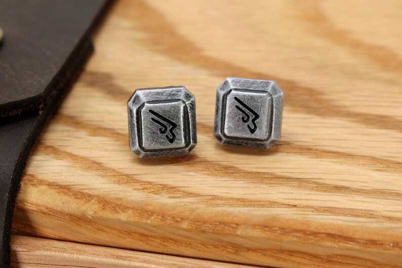 May include: Two silver cufflinks with a geometric design and a black symbol in the center. The cufflinks are on a wooden surface.
