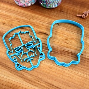 May include: Two blue plastic cookie cutters, one shaped like Frankenstein's face and the other shaped like a head.