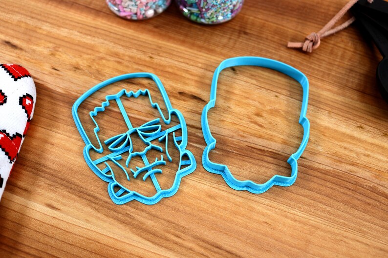 May include: Two blue plastic cookie cutters, one shaped like Frankenstein's face and the other shaped like a head.