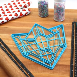 Puede incluir: Un cortador de galletas de pl&aacute;stico azul con forma de estrella geom&eacute;trica, un rodillo de madera, una regla negra, una esp&aacute;tula azul y dos frascos de espolvorear sobre una superficie de madera.