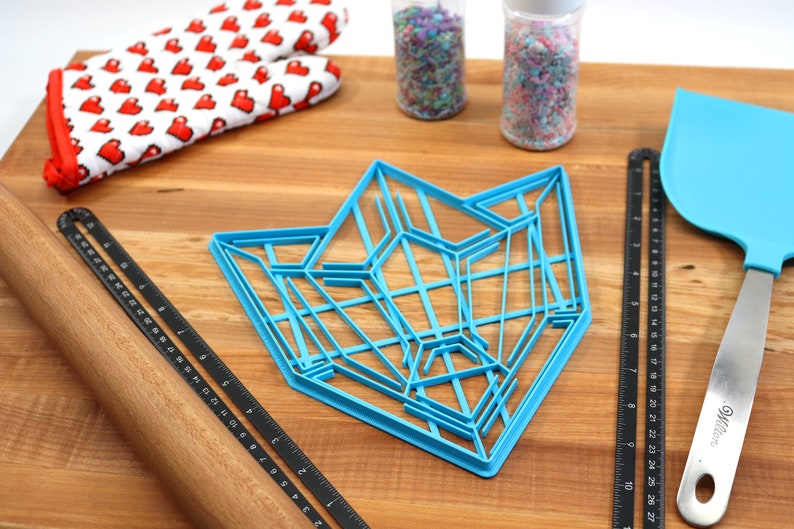 Puede incluir: Un cortador de galletas de pl&aacute;stico azul con forma de estrella geom&eacute;trica, un rodillo de madera, una regla negra, una esp&aacute;tula azul y dos frascos de espolvorear sobre una superficie de madera.
