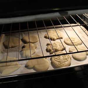 May include: A baking sheet with a variety of unbaked cookies in an oven. The cookies are shaped like different characters and are ready to be baked.