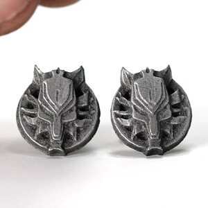 Op de afbeelding: Een paar zilverkleurige metalen oorbellen met een wolfskopontwerp. De wolfskop is omgeven door een cirkelvormig ontwerp met een getextureerd oppervlak.