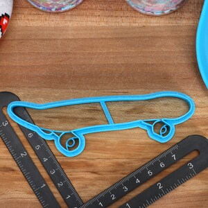 May include: A blue plastic cookie cutter shaped like a skateboard. The cutter is on a wooden surface with a ruler and a blue plate.