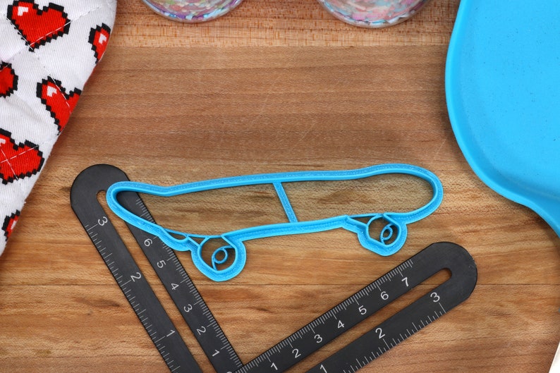 May include: A blue plastic cookie cutter shaped like a skateboard. The cutter is on a wooden surface with a ruler and a blue plate.