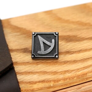 May include: A silver metal pin with a black background and a stylized silver symbol. The pin is square with rounded corners and has four small silver studs around the perimeter.