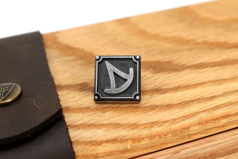 May include: A silver metal pin with a black background and a stylized silver symbol. The pin is square with rounded corners and has four small silver studs around the perimeter.