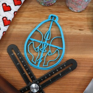 May include: A blue plastic cookie cutter shaped like an egg with a design of three birds inside. The cutter is on a wooden surface with a ruler and a spatula.