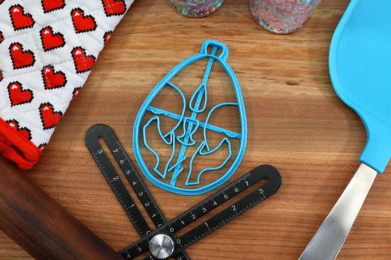 May include: A blue plastic cookie cutter shaped like an egg with a design of three birds inside. The cutter is on a wooden surface with a ruler and a spatula.