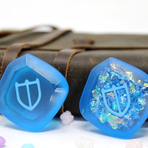 May include: Two blue resin dice with a shield symbol. One die has a smooth surface and the other has iridescent glitter embedded in the resin.