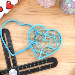 May include: Two blue plastic heart-shaped cookie cutters on a wooden surface. One cutter is a simple heart shape, while the other has a more intricate design with multiple smaller hearts inside.