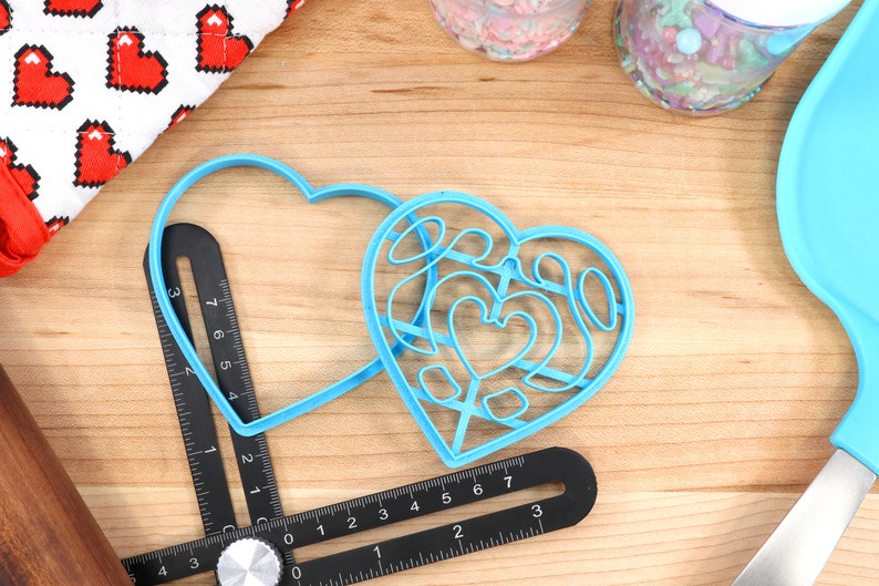 May include: Two blue plastic heart-shaped cookie cutters on a wooden surface. One cutter is a simple heart shape, while the other has a more intricate design with multiple smaller hearts inside.