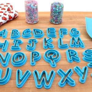 Op de afbeelding: Een set van 26 blauwe plastic alfabetkoekjesvormen op een houten oppervlak. De vormen zijn alfabetisch gerangschikt.
