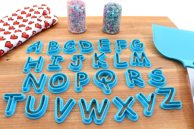 Op de afbeelding: Een set van 26 blauwe plastic alfabetkoekjesvormen op een houten oppervlak. De vormen zijn alfabetisch gerangschikt.