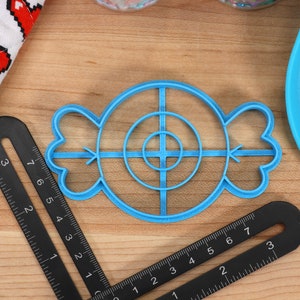 May include: A blue plastic cookie cutter shaped like a candy with a target design in the center. The cutter has a circle with four lines extending from the center.
