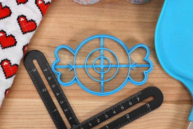 May include: A blue plastic cookie cutter shaped like a candy with a target design in the center. The cutter has a circle with four lines extending from the center.