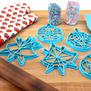 May include: A set of five blue plastic cookie cutters shaped like ornaments, a red and white oven mitt, a wooden rolling pin, and two jars of sprinkles on a wooden cutting board.
