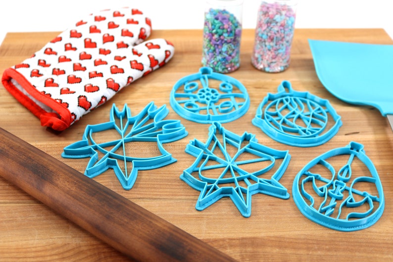 May include: A set of five blue plastic cookie cutters shaped like ornaments, a red and white oven mitt, a wooden rolling pin, and two jars of sprinkles on a wooden cutting board.
