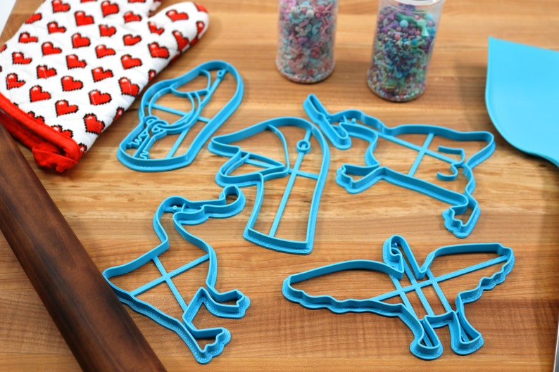 May include: A set of blue plastic cookie cutters shaped like a duck, a bird, a hat, a shoe, and a unicorn. The cutters are on a wooden surface.