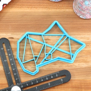 May include: A light blue plastic cookie cutter shaped like a geometric design. The cutter is on a wooden surface.