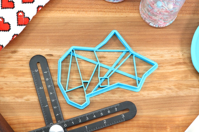 May include: A light blue plastic cookie cutter shaped like a geometric design. The cutter is on a wooden surface.
