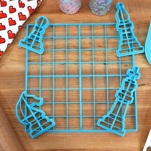 May include: A blue plastic grid with four blue plastic chess piece cookie cutters. The cutters are shaped like a knight, bishop, rook, and king. The grid is on a wooden surface.