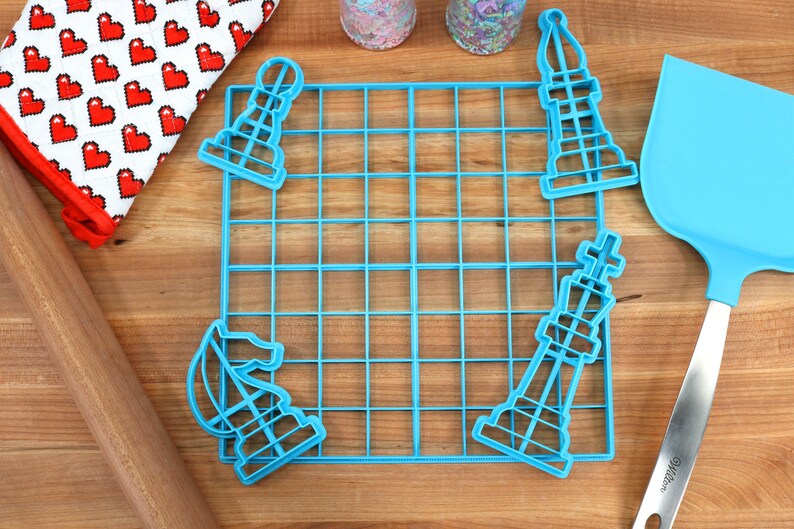 May include: A blue plastic grid with four blue plastic chess piece cookie cutters. The cutters are shaped like a knight, bishop, rook, and king. The grid is on a wooden surface.