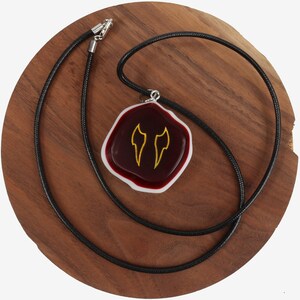 Op de afbeelding: Een rode en witte glazen hanger met een gouden ontwerp van twee vleugels hangt aan een zwarte koordsnoer. De ketting wordt getoond op een houten oppervlak.