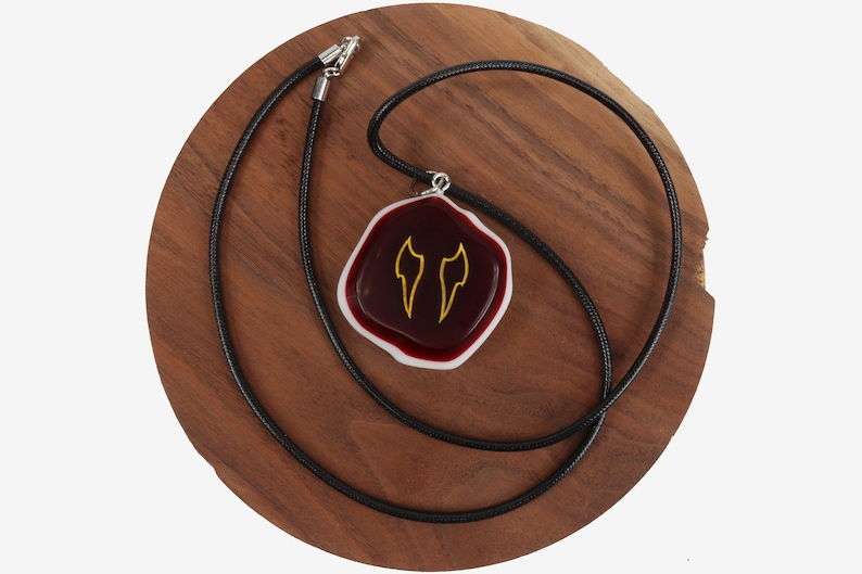 Op de afbeelding: Een rode en witte glazen hanger met een gouden ontwerp van twee vleugels hangt aan een zwarte koordsnoer. De ketting wordt getoond op een houten oppervlak.