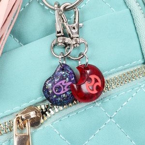 Puede incluir: Una mochila acolchada de color turquesa con un borla rosa y dos abalorios unidos a un llavero plateado. Un abalorio es un coraz&oacute;n rojo con un s&iacute;mbolo rosa, y el otro es un coraz&oacute;n azul con un s&iacute;mbolo morado.