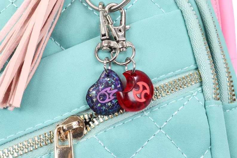 Puede incluir: Una mochila acolchada de color turquesa con un borla rosa y dos abalorios unidos a un llavero plateado. Un abalorio es un coraz&oacute;n rojo con un s&iacute;mbolo rosa, y el otro es un coraz&oacute;n azul con un s&iacute;mbolo morado.