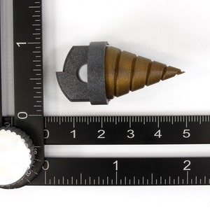 May include: A brown and gray drill bit with a spiral design, approximately 2 inches long, is positioned on a white surface. The drill bit is next to a black ruler with inch markings. The drill bit is a tool used for drilling holes.
