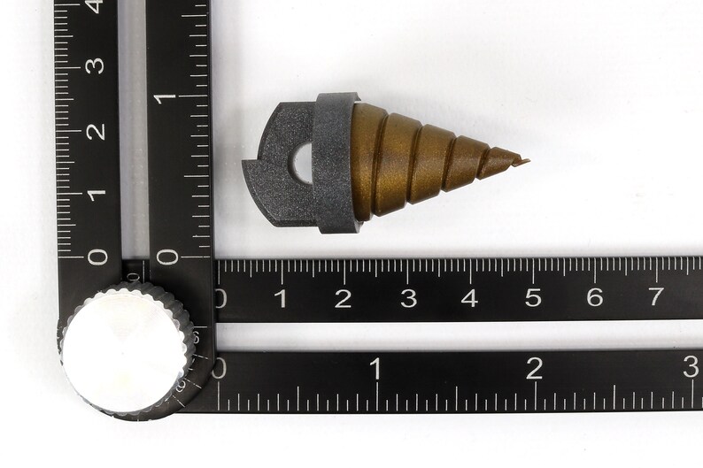 May include: A brown and gray drill bit with a spiral design, approximately 2 inches long, is positioned on a white surface. The drill bit is next to a black ruler with inch markings. The drill bit is a tool used for drilling holes.